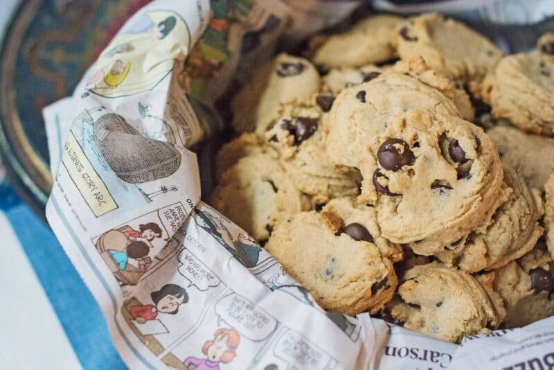A basket lined with comic newspaper pages holds a pile of delicious Chocolate Chip Cookies, displayed enticingly on a blue and white surface.