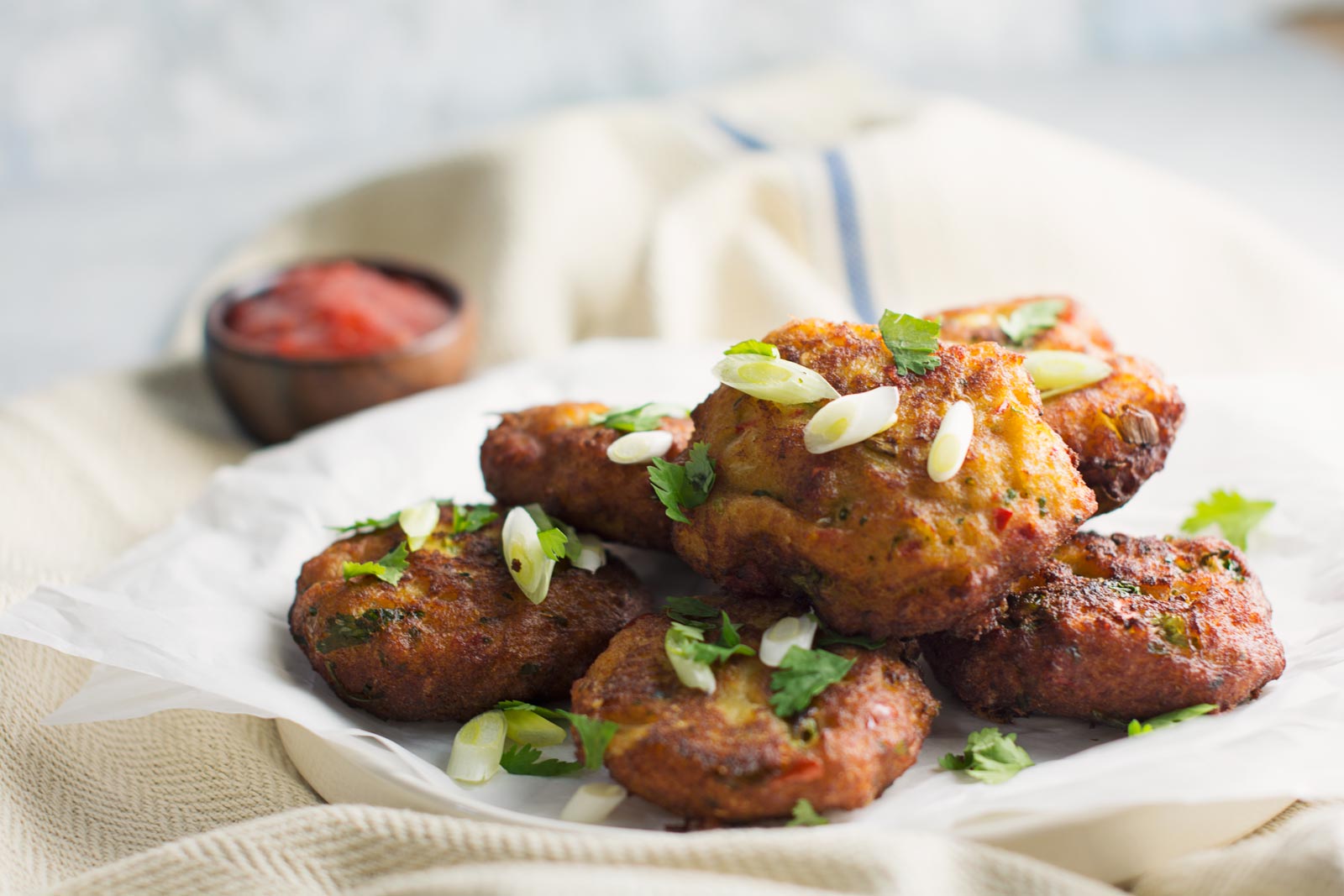A plate of crispy vegetable fritters and savory fish cakes, garnished with chopped green onions and cilantro, served on parchment paper with a small bowl of red dipping sauce in the background.