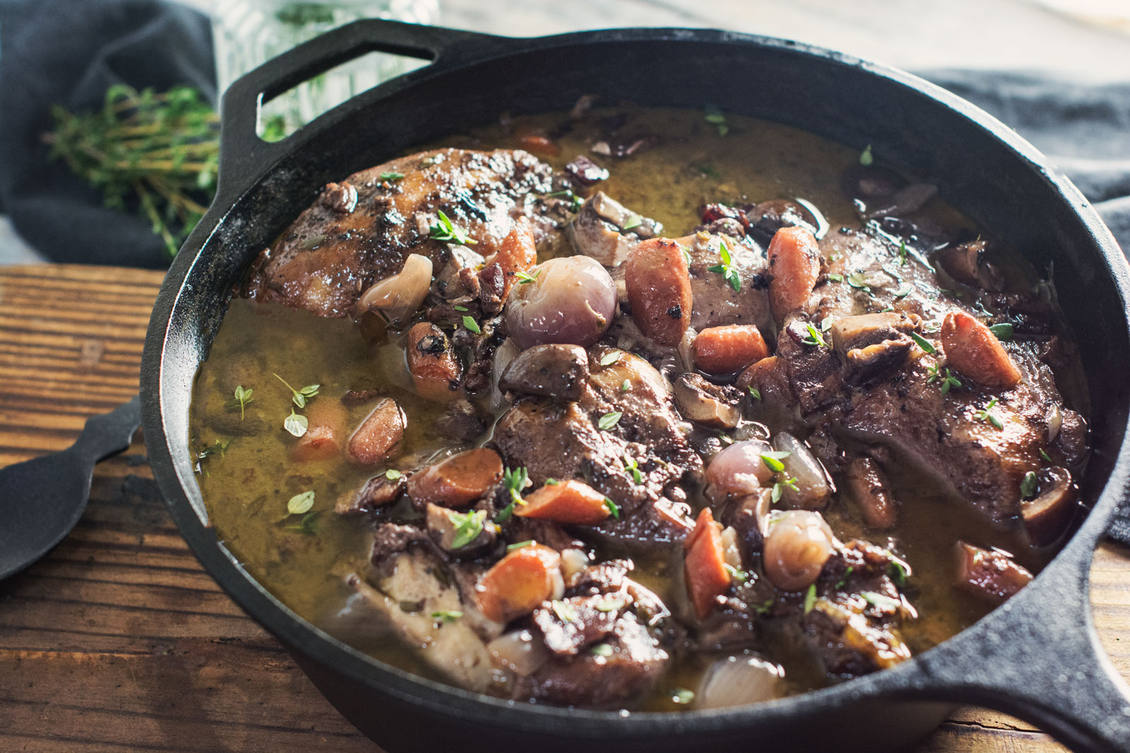 A cast iron skillet filled with Coq au Vin—braised chicken, carrots, pearl onions, and herbs in a rich, brown sauce—sits on a wooden table next to a bundle of fresh thyme.