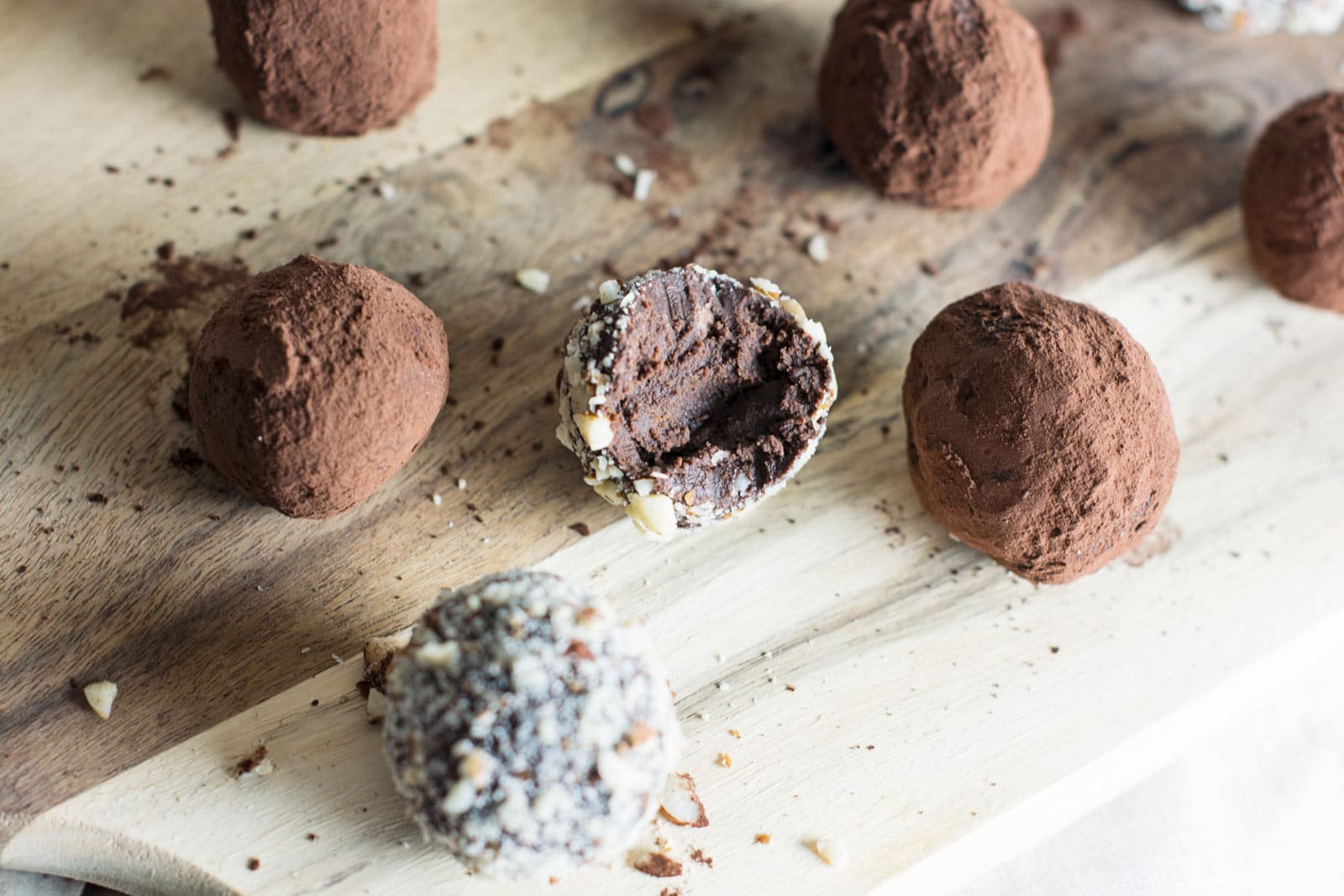 Several chocolate truffles, some coated in cocoa powder and others in chopped nuts, rest on a wooden board. One chocolate truffle with a nut coating is bitten open, revealing a rich, dense chocolate center.