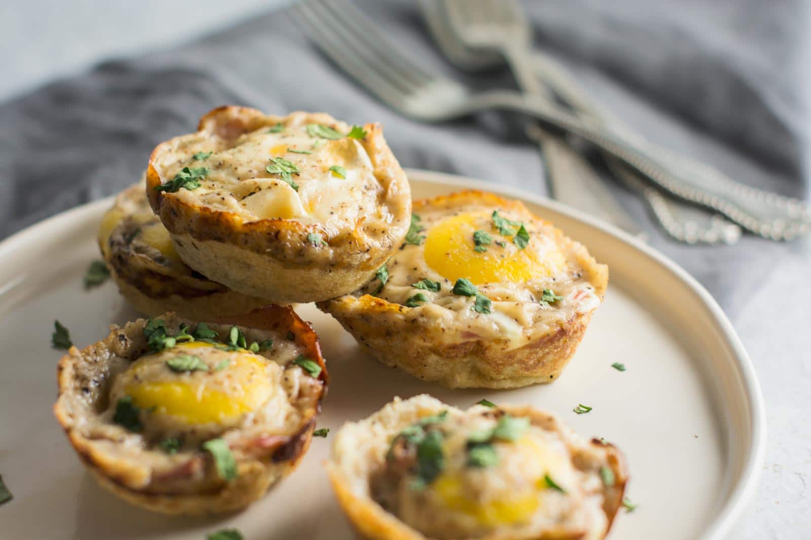 Four savory Breakfast Bites muffin cups with eggs, herbs, and a golden-brown crust are served on a white plate, with forks and a gray napkin in the background.