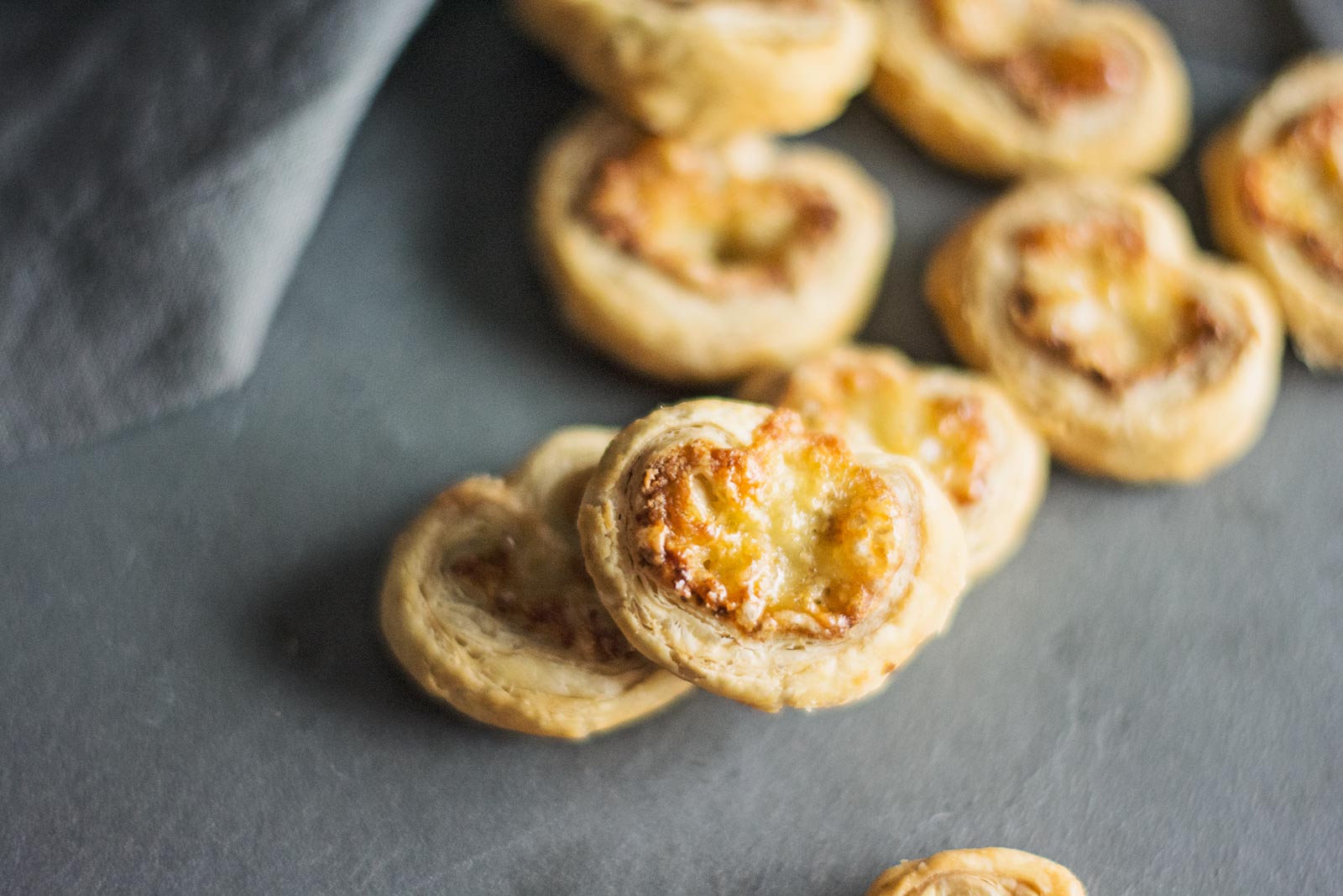 A close-up of several golden, flaky cheese palmiers arranged on a dark surface, with a gray cloth partially visible in the background.