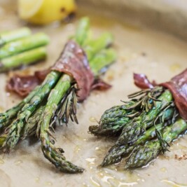 Three bundles of roasted asparagus spears wrapped in prosciutto rest on parchment paper, lightly drizzled with oil. A wedge of lemon is visible in the background, complementing the savory roasted asparagus.