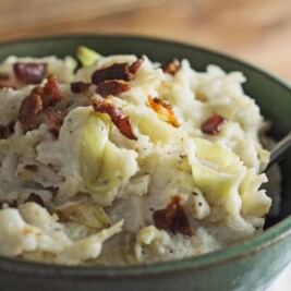 A bowl of creamy Colcannon—mashed potatoes blended with cabbage and topped with crispy bacon pieces—rests invitingly, a spoon nestled inside.