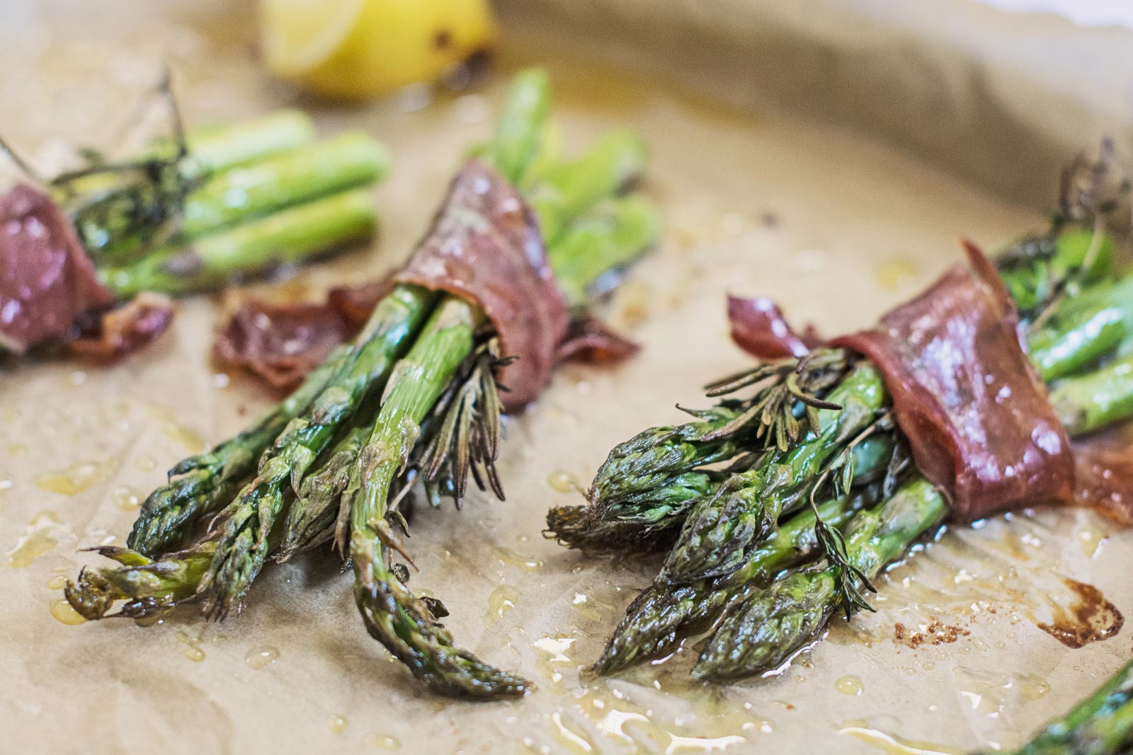 Three bundles of roasted asparagus spears wrapped in prosciutto rest on parchment paper, lightly drizzled with oil. A wedge of lemon is visible in the background, complementing the savory roasted asparagus.