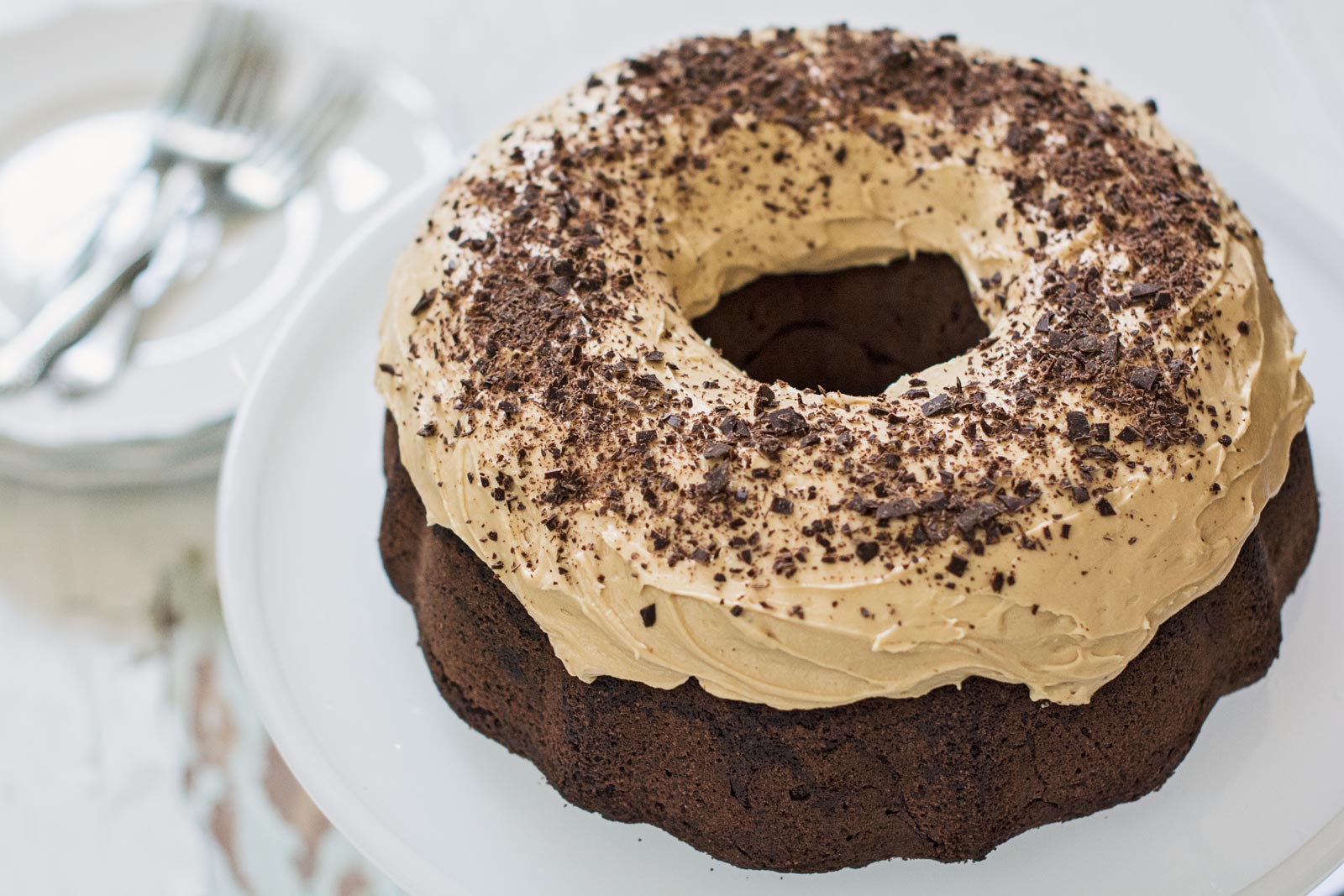 A Cardamom Chocolate Cake bundt topped with creamy light brown frosting and sprinkled with chocolate shavings, displayed on a white cake stand. Plates and forks are blurred in the background.