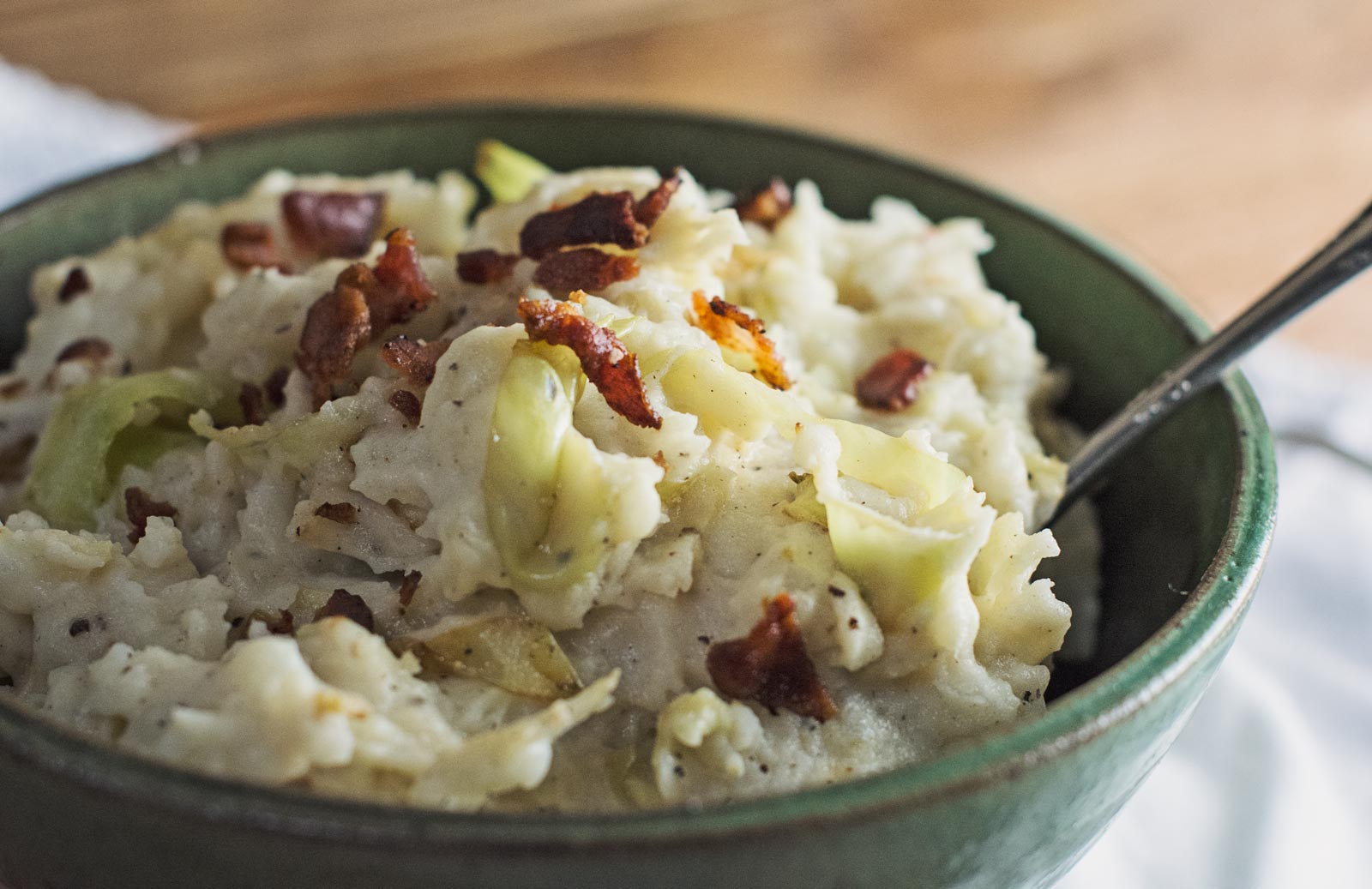 A bowl of creamy Colcannon—mashed potatoes blended with cabbage and topped with crispy bacon pieces—rests invitingly, a spoon nestled inside.
