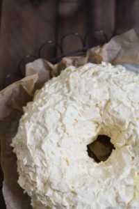A close-up of a Pea Pickers Cake bundt, covered in thick, textured white frosting, set on brown parchment paper with part of a wire rack visible in the background.