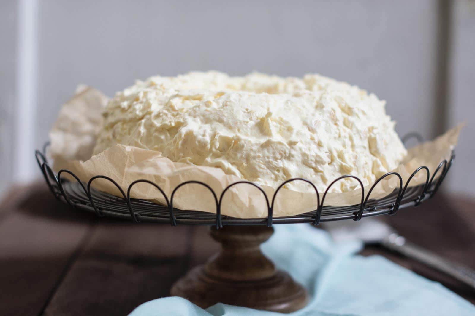 A Pea Pickers Cake with rough, textured white icing sits on a parchment-lined wire cake stand with a wooden base, placed on a wooden surface with a light blue cloth nearby.