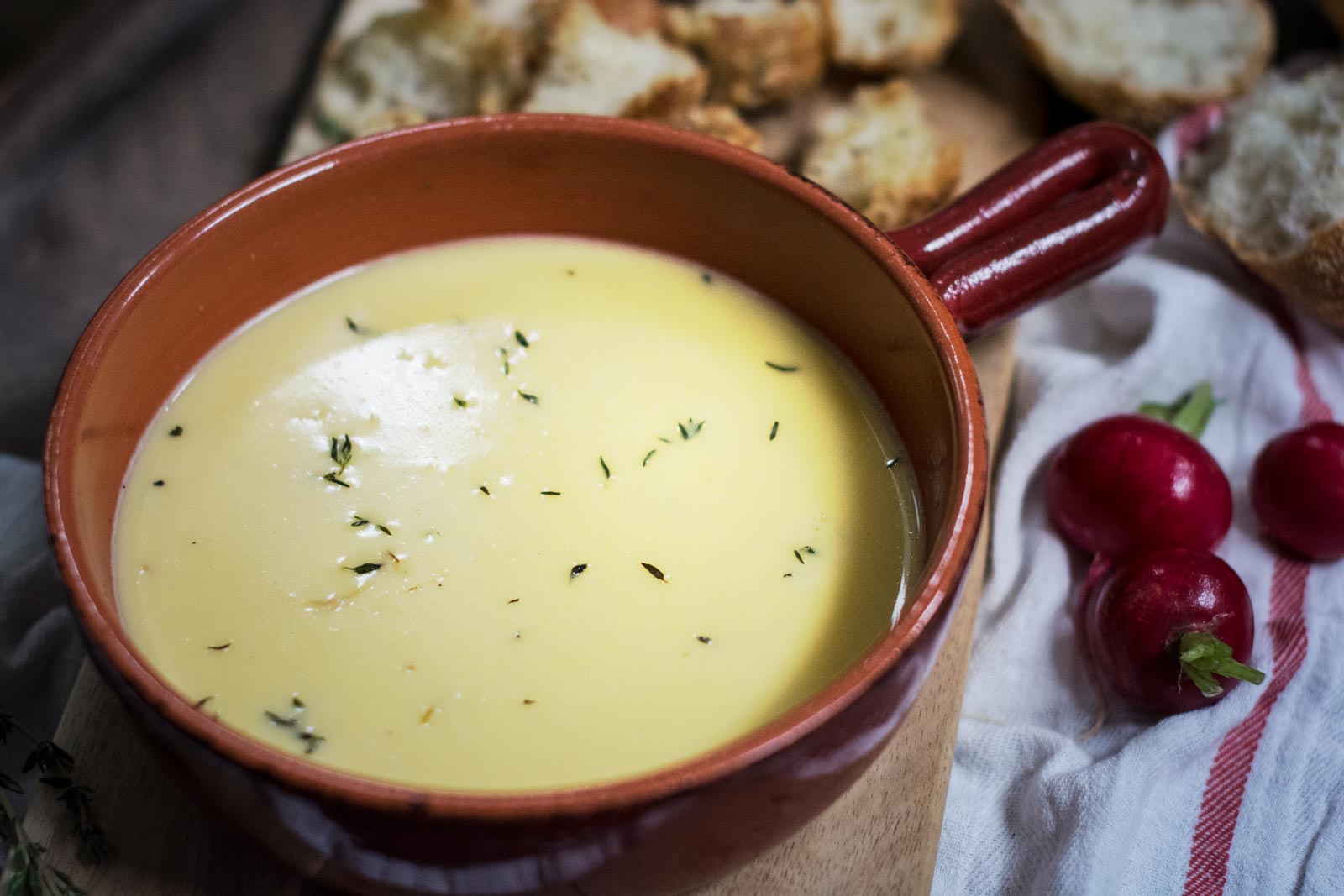 A brown ceramic bowl filled with creamy yellow cheese fondue, garnished with fresh herbs, sits on a wooden board. Slices of bread and two red radishes rest nearby on a white and red striped cloth.
