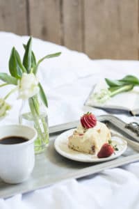 A breakfast tray with a cup of coffee, strawberry sweet rolls topped with fresh berries, and a small vase of white flowers, set on a bed with a white blanket and an open notebook in the background.