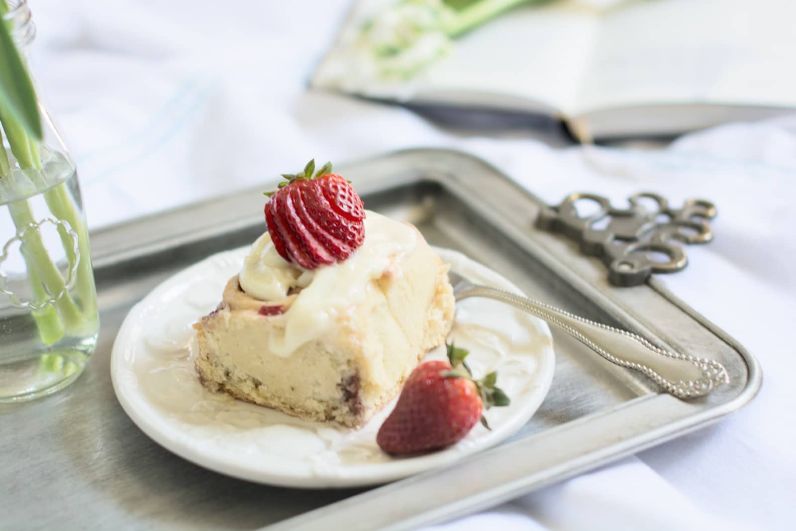 A slice of cheesecake topped with a glazed strawberry and cream sits on a white plate with a vintage fork, placed on a silver tray; beside it, Strawberry Sweet Rolls and an open book are blurred in the background.