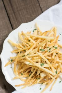 A white plate filled with thin, crispy fries garnished with chopped parsley sits on a wooden surface, accompanied by a white cloth nearby.