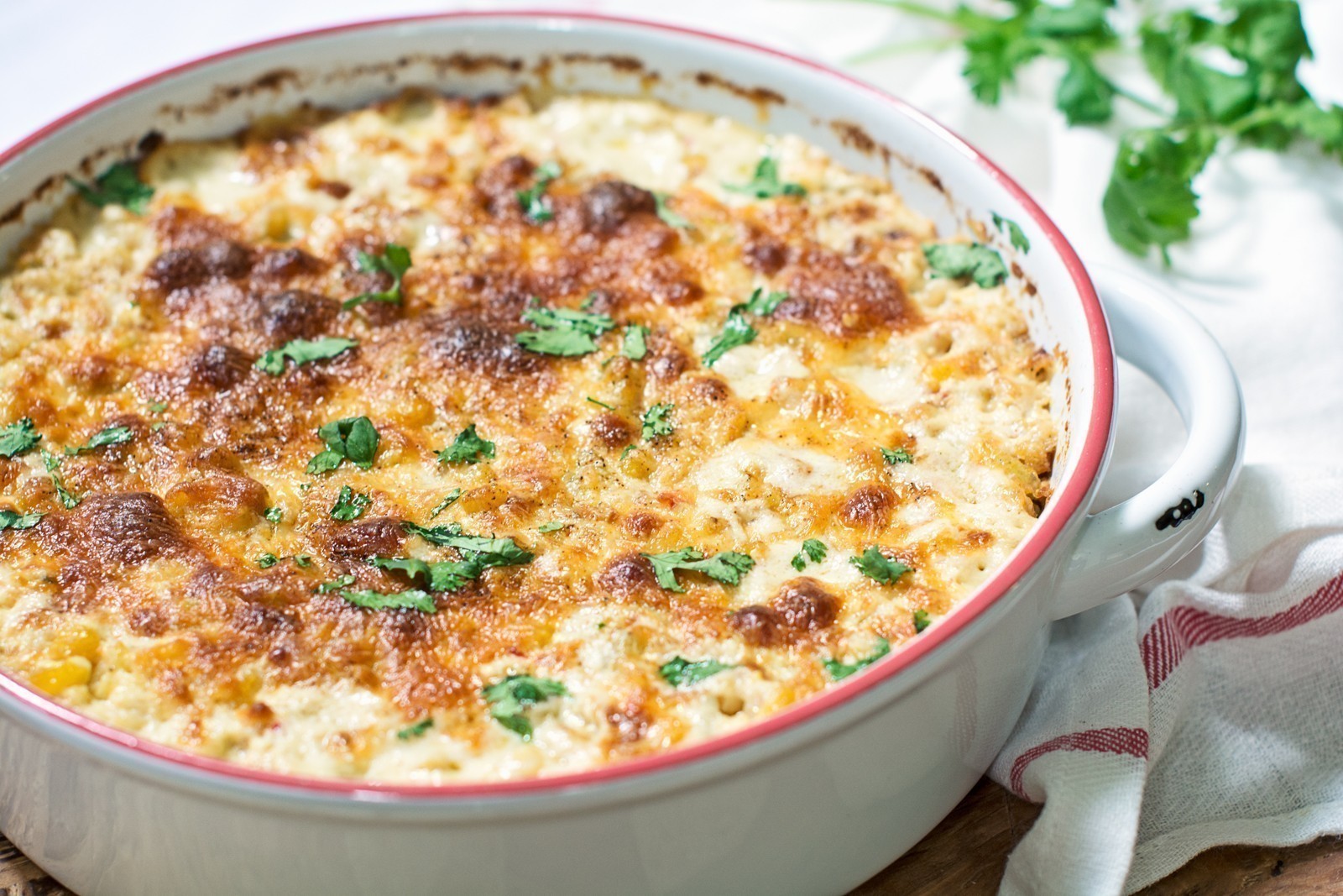 A baked cheesy corn dip casserole topped with golden brown melted cheese and sprinkled with fresh herbs sits in a round white baking dish, with a white towel and green herbs in the background.