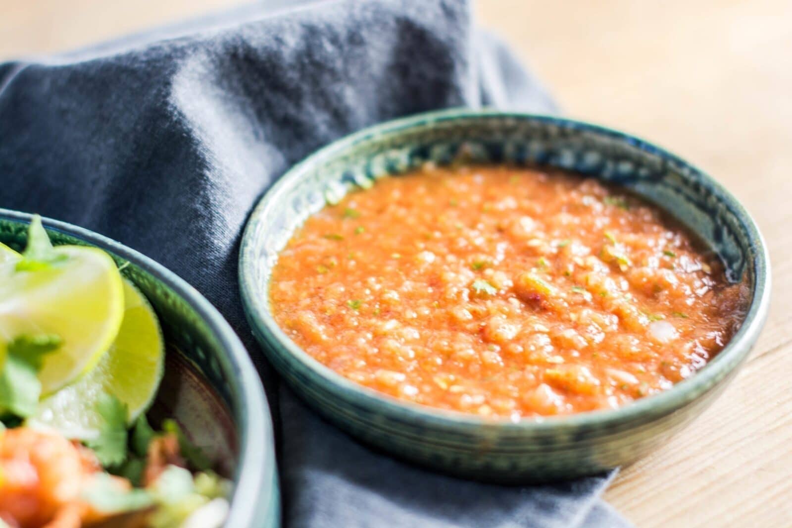 A bowl of chunky Chipotle Salsa sits on a blue cloth next to a plate with lime wedges and fresh greens on a wooden surface.