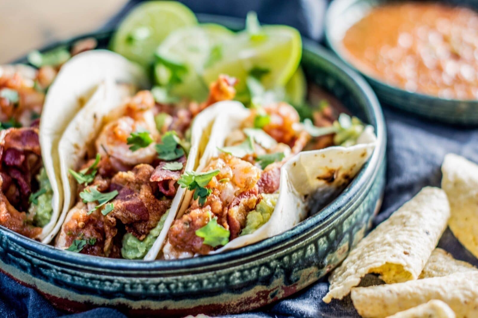 Three soft shrimp tacos filled with beans, vegetables, and fresh cilantro are served in a rustic bowl with lime wedges. A side of salsa and a few tortilla chips are visible in the background.