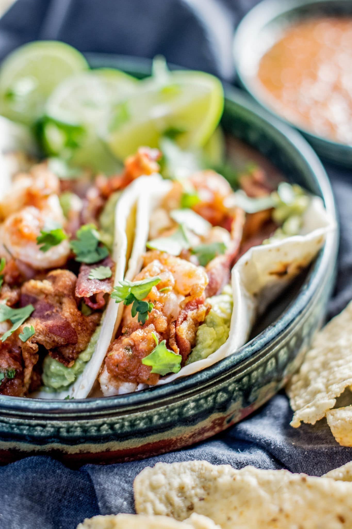 Three Shrimp Tacos filled with guacamole, salsa, and cilantro are served in a green ceramic dish with lime wedges. A side of tortilla chips and a bowl of salsa add the perfect touch in the background.