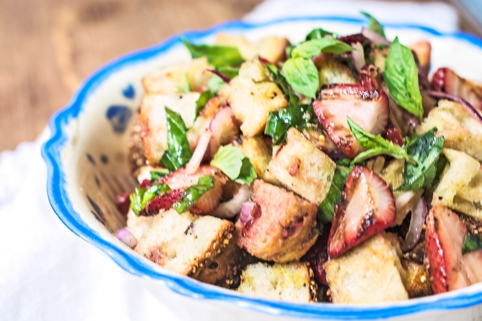 A close-up of a Strawberry Panzanella in a blue-rimmed bowl, featuring toasted bread cubes, sliced strawberries, fresh basil leaves, and thinly sliced red onions.