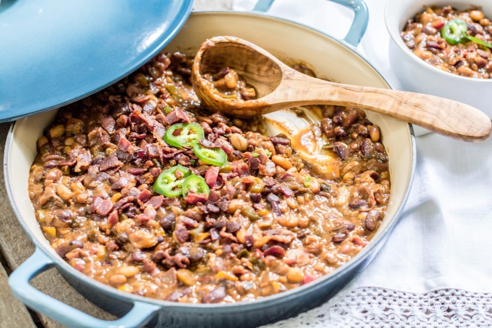 A pot of smoked bbq beans topped with chopped bacon and sliced jalapeños, with a wooden spoon resting inside. A small serving is in a bowl nearby, and the pot’s light blue lid is partially open.