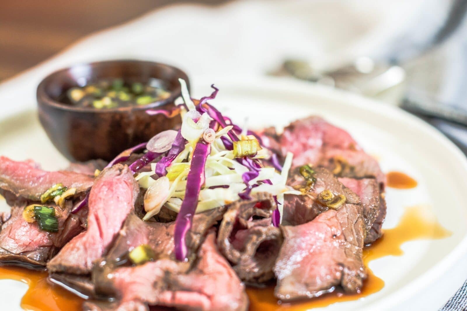 Sliced medium-rare Beef Tataki is arranged on a plate, topped with shredded cabbage and red cabbage, and garnished with green onions. A small bowl of dipping sauce is placed in the background.