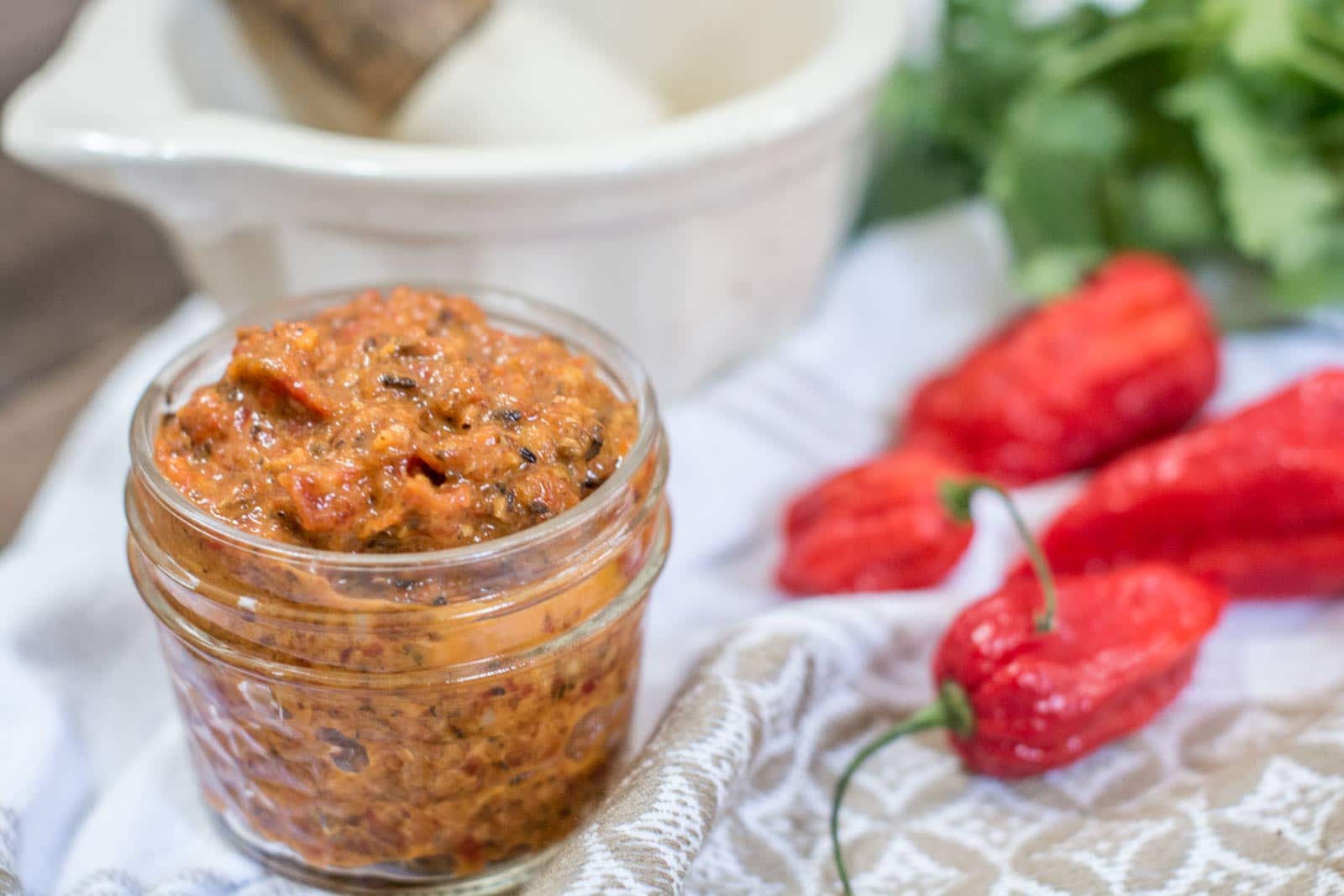 A small glass jar filled with chunky red harissa chili paste sits on a cloth next to whole red chili peppers and fresh cilantro, with a white mortar and pestle in the background.