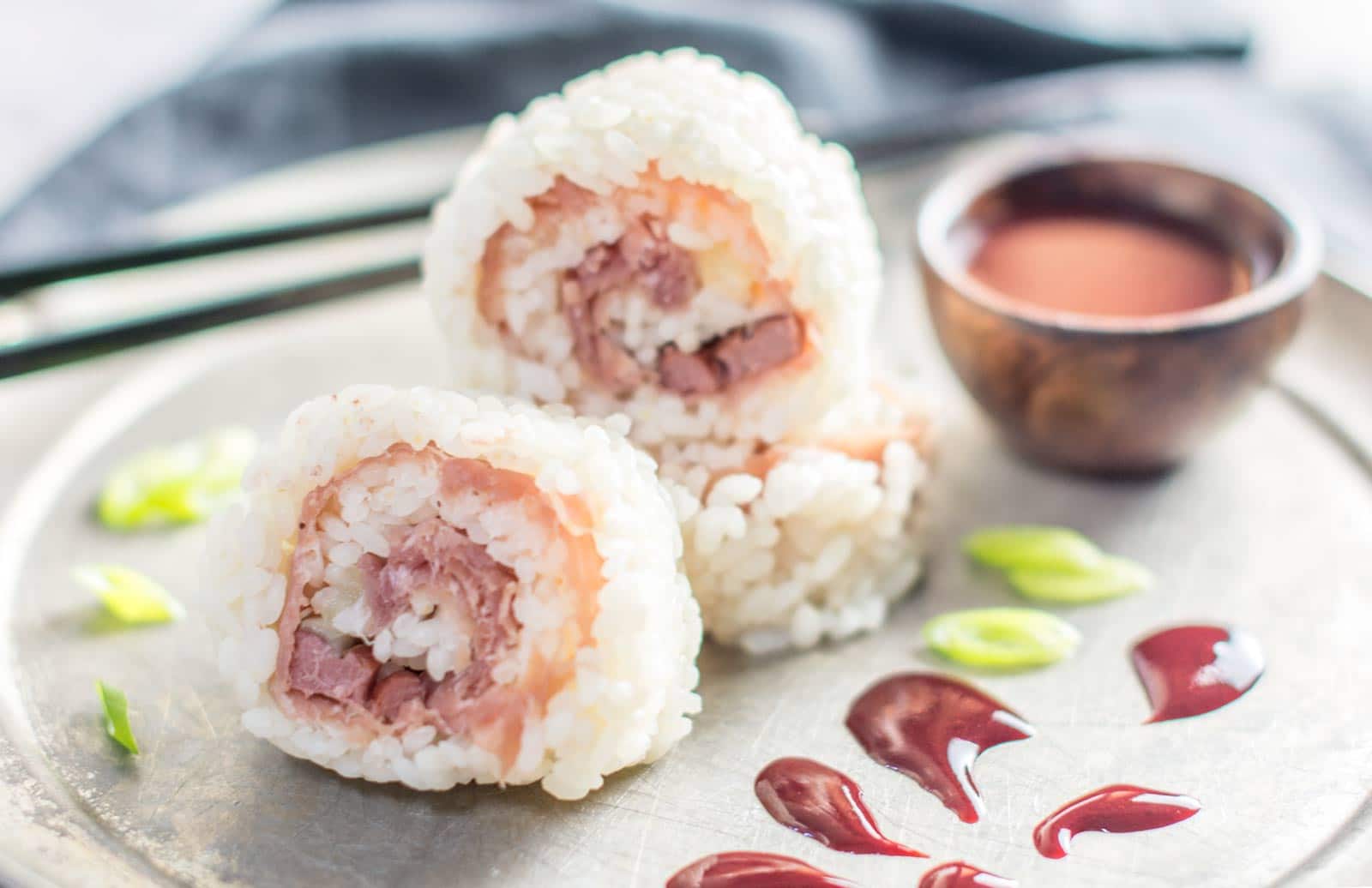 Two sushi rolls with rice on the outside and raw fish inside are on a plate, garnished with green onions. Dipping sauce in a small bowl and a decorative sauce pattern are also on the plate. Black chopsticks rest in the background.