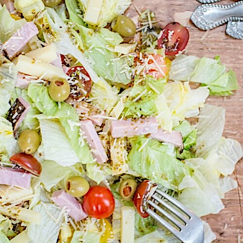 A close-up of a 1905 Salad with lettuce, cherry tomatoes, olives, ham strips, cheese, and grated parmesan, with a fork and part of a silver utensil visible on the right side.