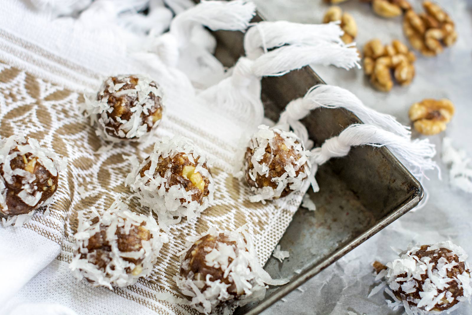 Energy bites made with dates and coated in shredded coconut rest on a patterned cloth beside a metal tray. Walnut pieces and shredded coconut are scattered around, adding to the rustic presentation.