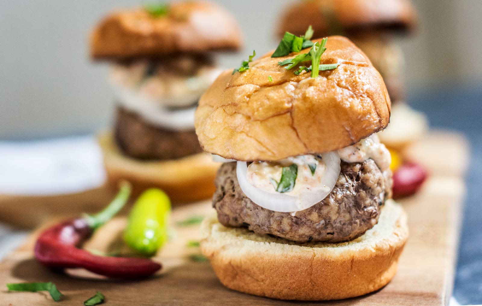 A close-up of a lamb burger slider with a soft bun, sliced onion, creamy sauce, and herbs on top, served on a wooden board with chili peppers and another slider in the background.