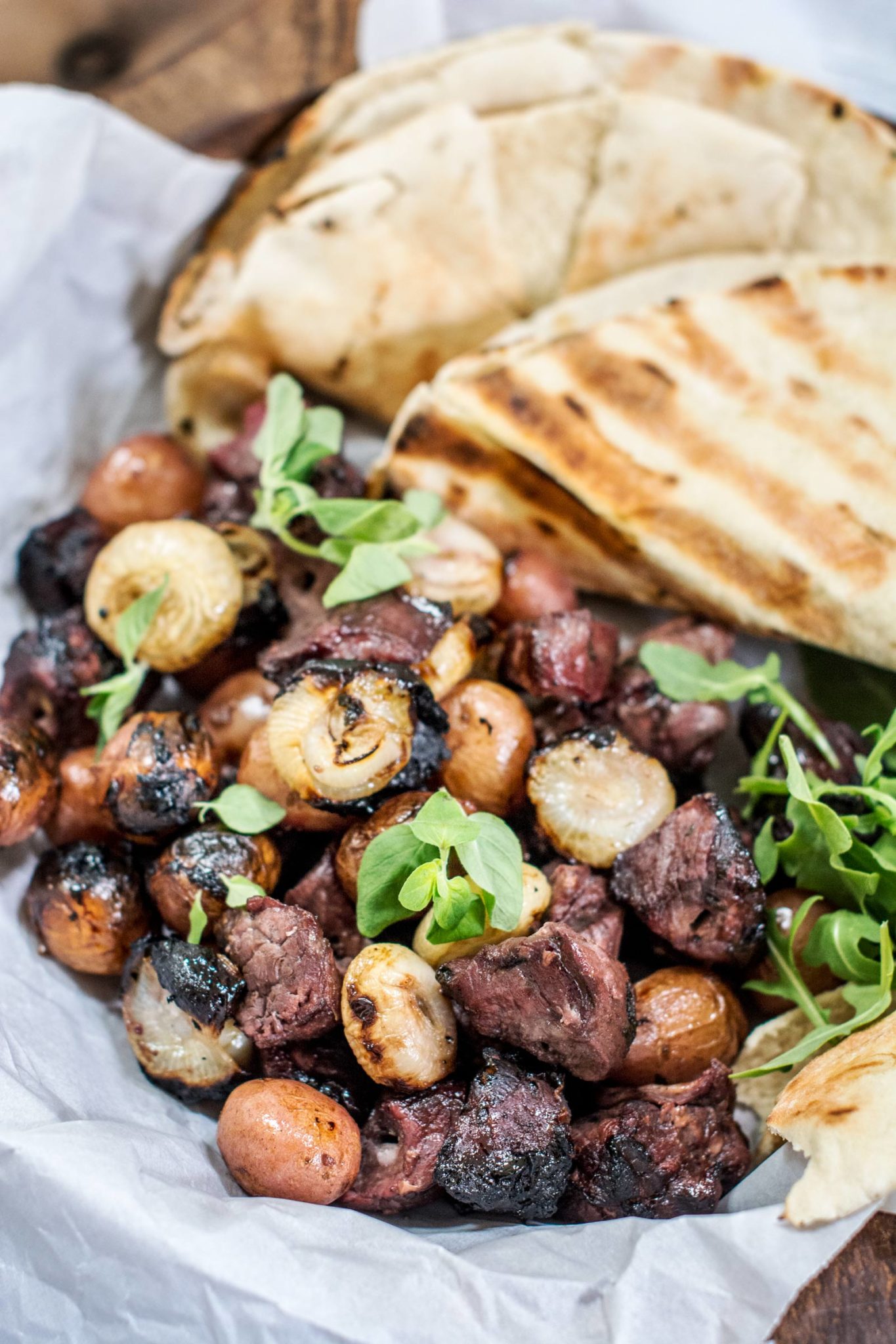 Honey Lamb Kabobs: Grilled skewers of marinated lamb and pearl onions served with warm pita bread, fresh greens, and herbs on parchment paper.