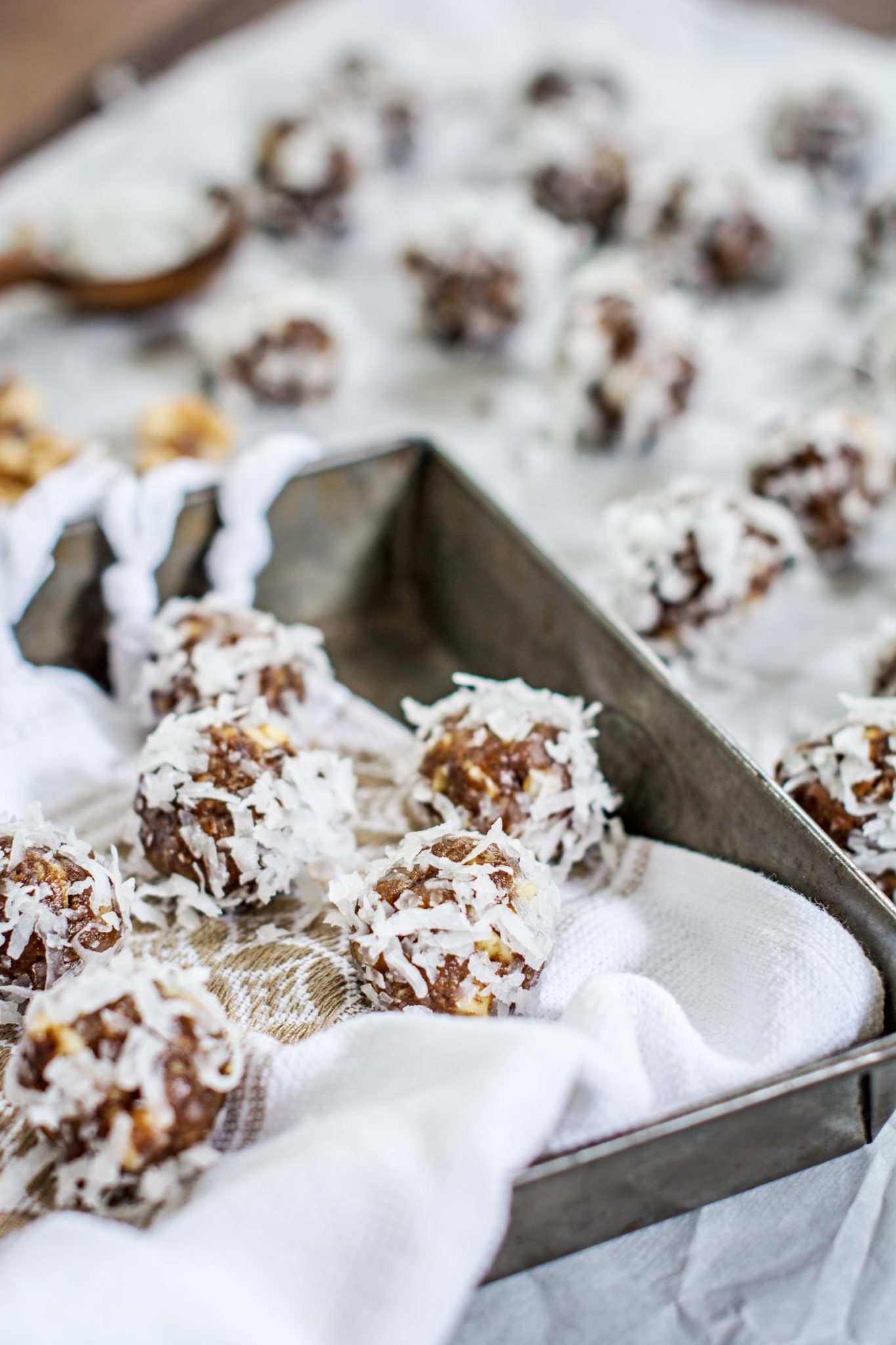 A tray and a table are lined with white cloth and topped with chocolate energy balls made from dates and coated in shredded coconut, with more energy balls blurred in the background.