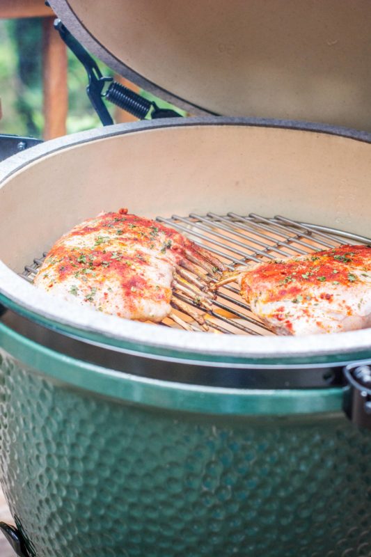 Two seasoned racks of ribs and tender lamb are cooking on a grill inside a green ceramic kamado-style smoker with the lid open.