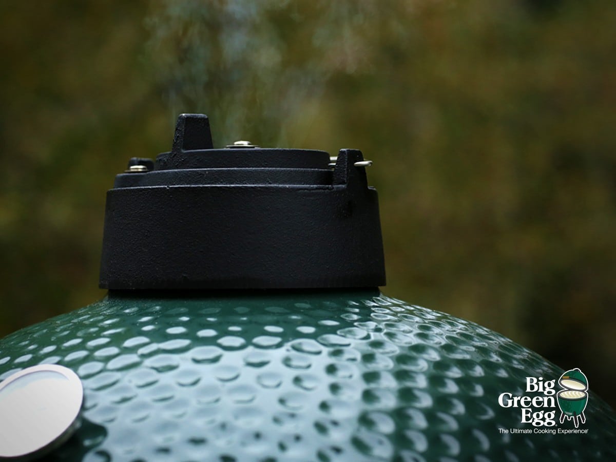Close-up of the top vent and green textured lid of a Big Green Egg ceramic grill, with steam rising as tender lamb cooks inside. The Big Green Egg logo is in the bottom right corner. Blurry outdoor background.
