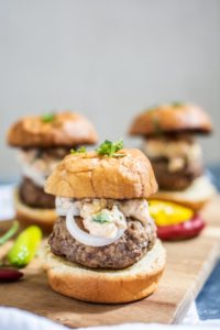 Three gourmet sliders with thick lamb burger patties, creamy topping, and onion slices on toasted buns, garnished with herbs, served on a wooden board with colorful sliced peppers in the background.