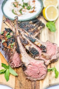 Grilled lamb chops with a pink center served on a wooden board, garnished with fresh mint. A bowl of yogurt sauce and lemon halves are in the background, highlighting the juicy lamb flavors.
