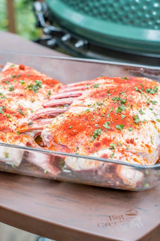 Two racks of lamb coated in spices and herbs sit in a glass dish on a wooden surface, with a green ceramic barbecue grill blurred in the background, ready for grilling perfection.