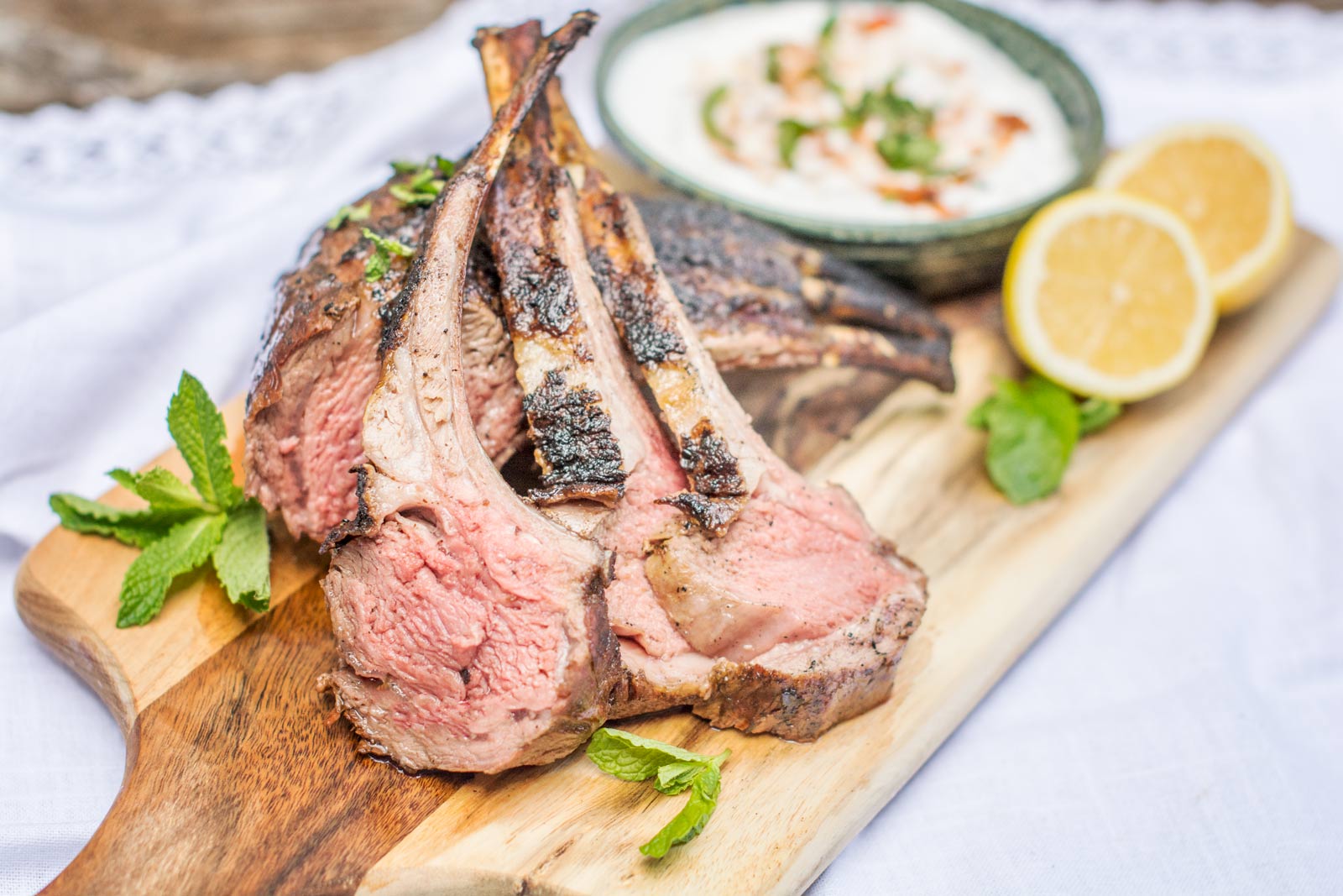 Grilled lamb chops arranged on a wooden board with fresh mint, lemon halves, and a bowl of creamy lamb dip garnished with herbs in the background.