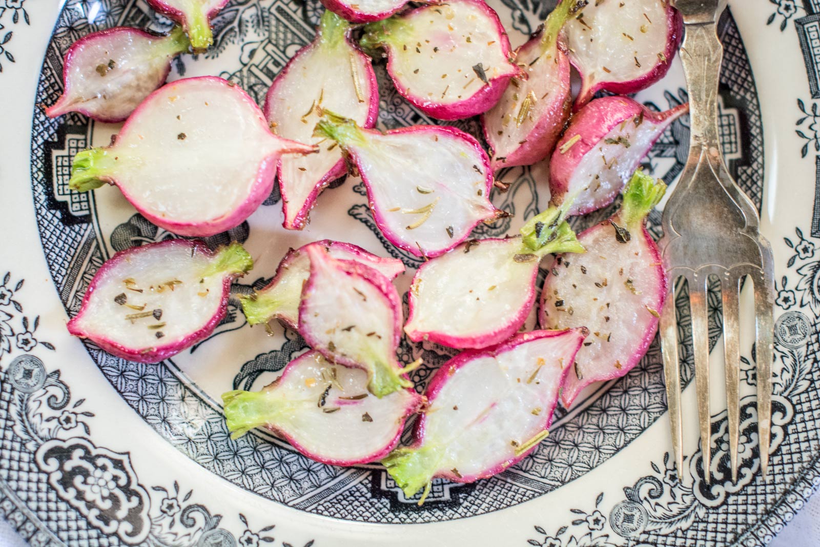 A plate with halved radishes sprinkled with herbs and pepper, showcasing the crisp freshness of radishes beside an ornate silver fork on a decorative black and white patterned dish.