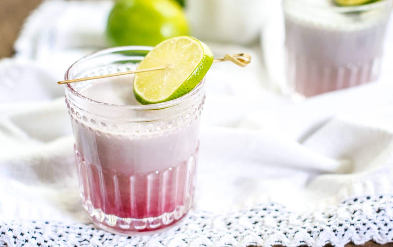A glass with a layered pink and white Coconut Passion Fruit drink, garnished with a lime slice on a cocktail pick, sits on a white cloth with a lace edge. Another similar glass and limes are blurred in the background.