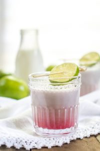 A glass of layered pink and white Coconut Passion Fruit drink, garnished with a slice of lime on the rim, set on a white lace cloth with blurred limes and a glass bottle in the background.