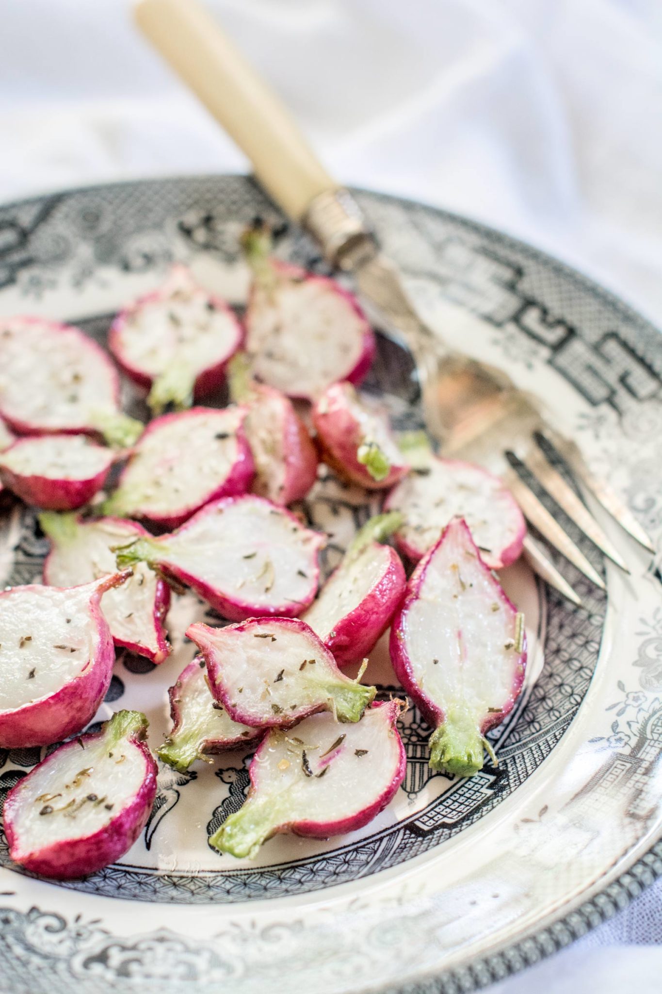 A decorative plate with roasted, halved radishes sprinkled with herbs sits alongside a vintage fork and knife, placed on a light-colored surface—perfect for showcasing the vibrant colors of fresh radishes.