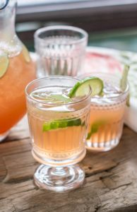 Two glasses of a light pink Scarlett OHara Drink garnished with lime wedges sit on a rustic wooden surface, with a pitcher and more Southern Comfort Cocktails in the background.