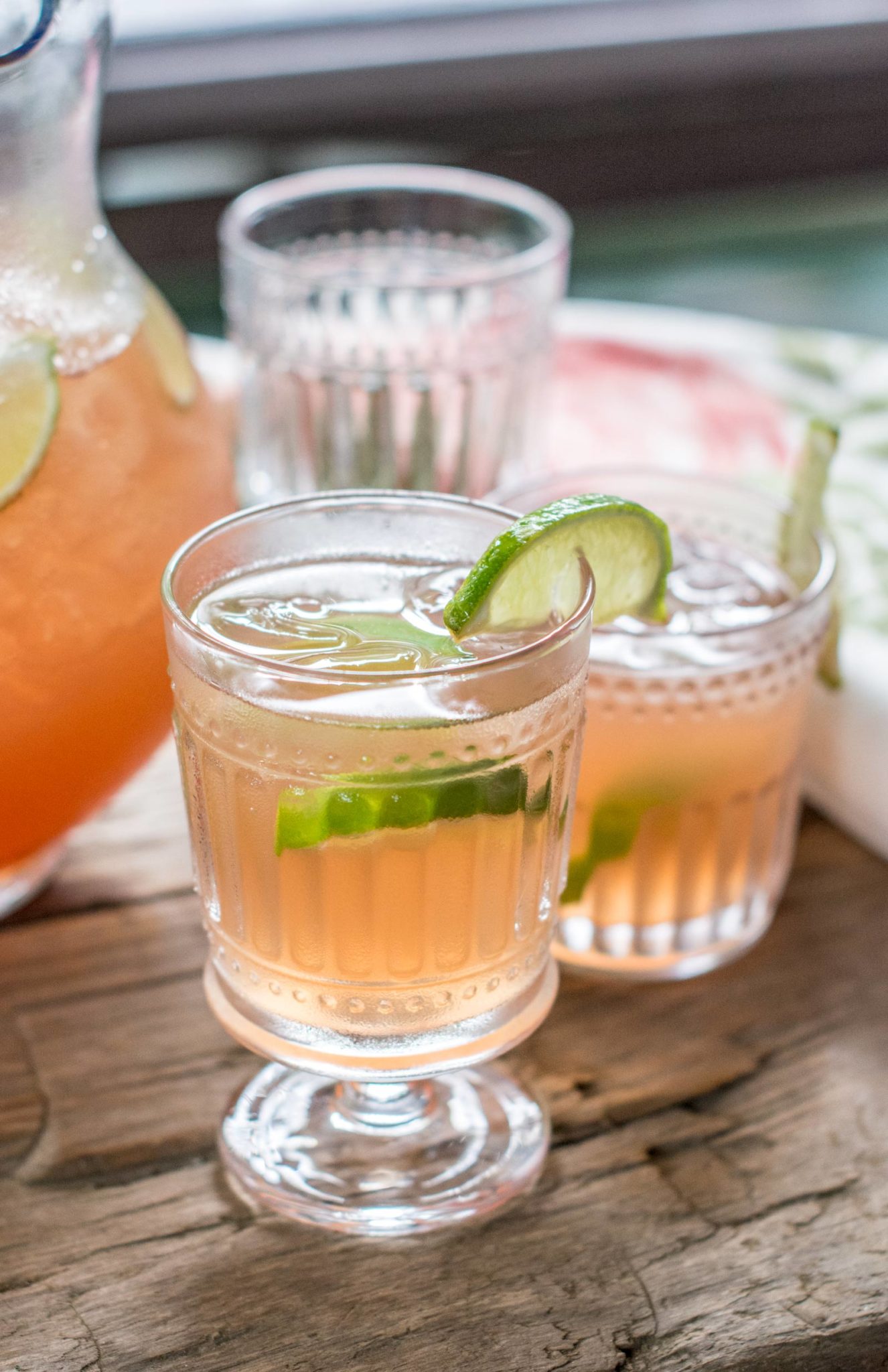 Two glasses of a light pink Scarlett OHara Drink garnished with lime wedges sit on a rustic wooden surface, with a pitcher and more Southern Comfort Cocktails in the background.