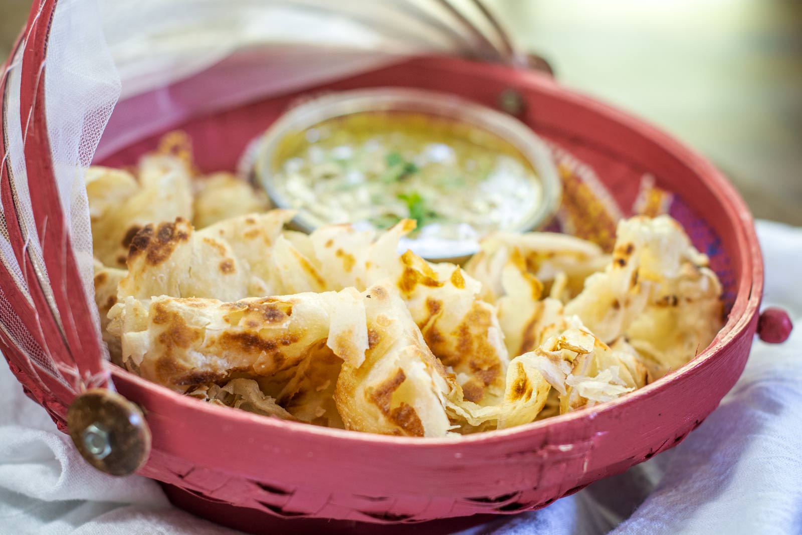 A red basket lined with white cloth holds pieces of flaky, golden-brown Roti Canai with a small bowl of curry or dipping sauce in the center. A transparent mesh cover is partially open above the basket.