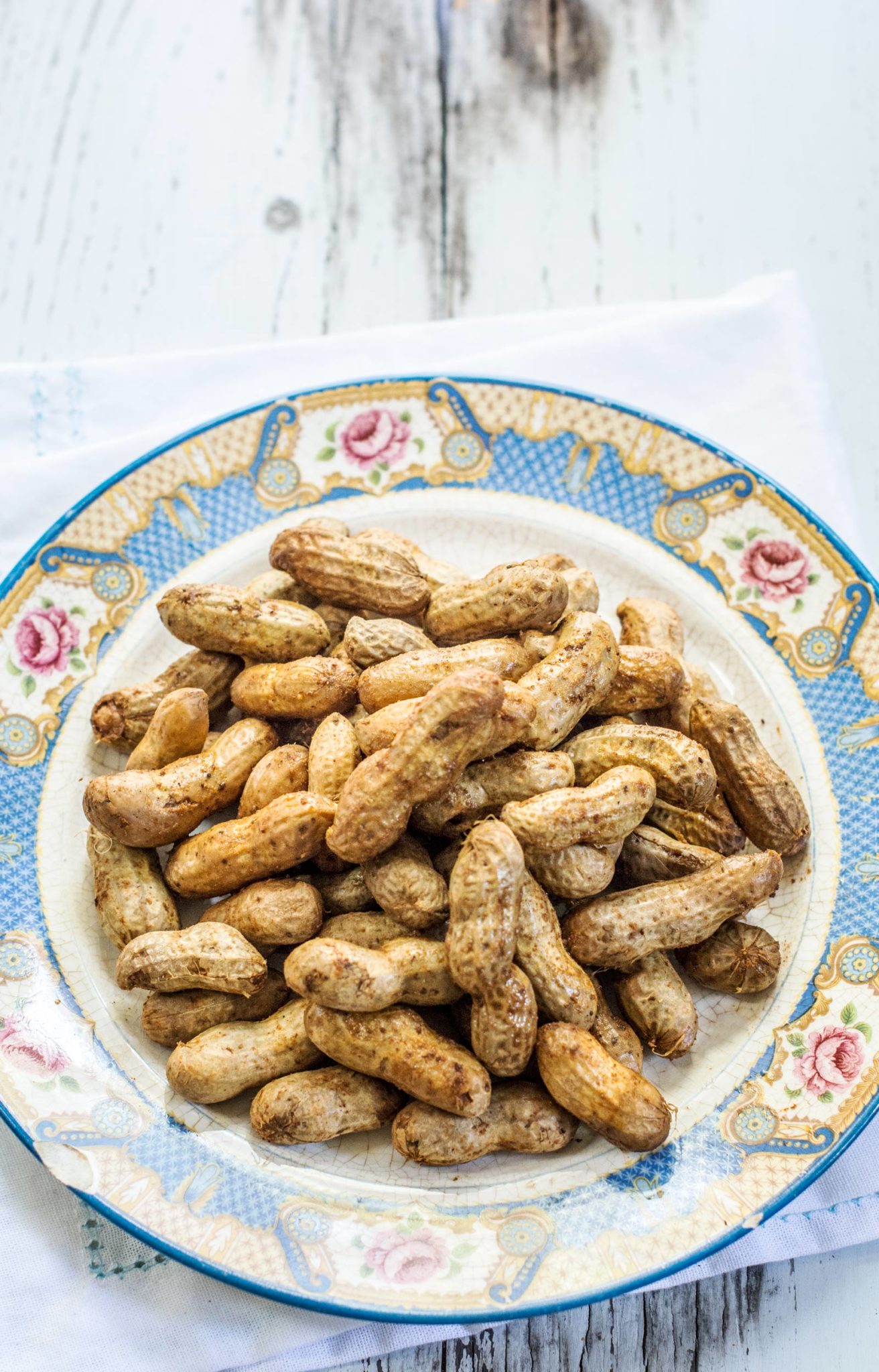 A decorative plate with a floral pattern holds a pile of boiled peanuts in their shells, placed on a white napkin over a rustic wooden surface—perfect for enjoying classic Southern boiled peanuts.