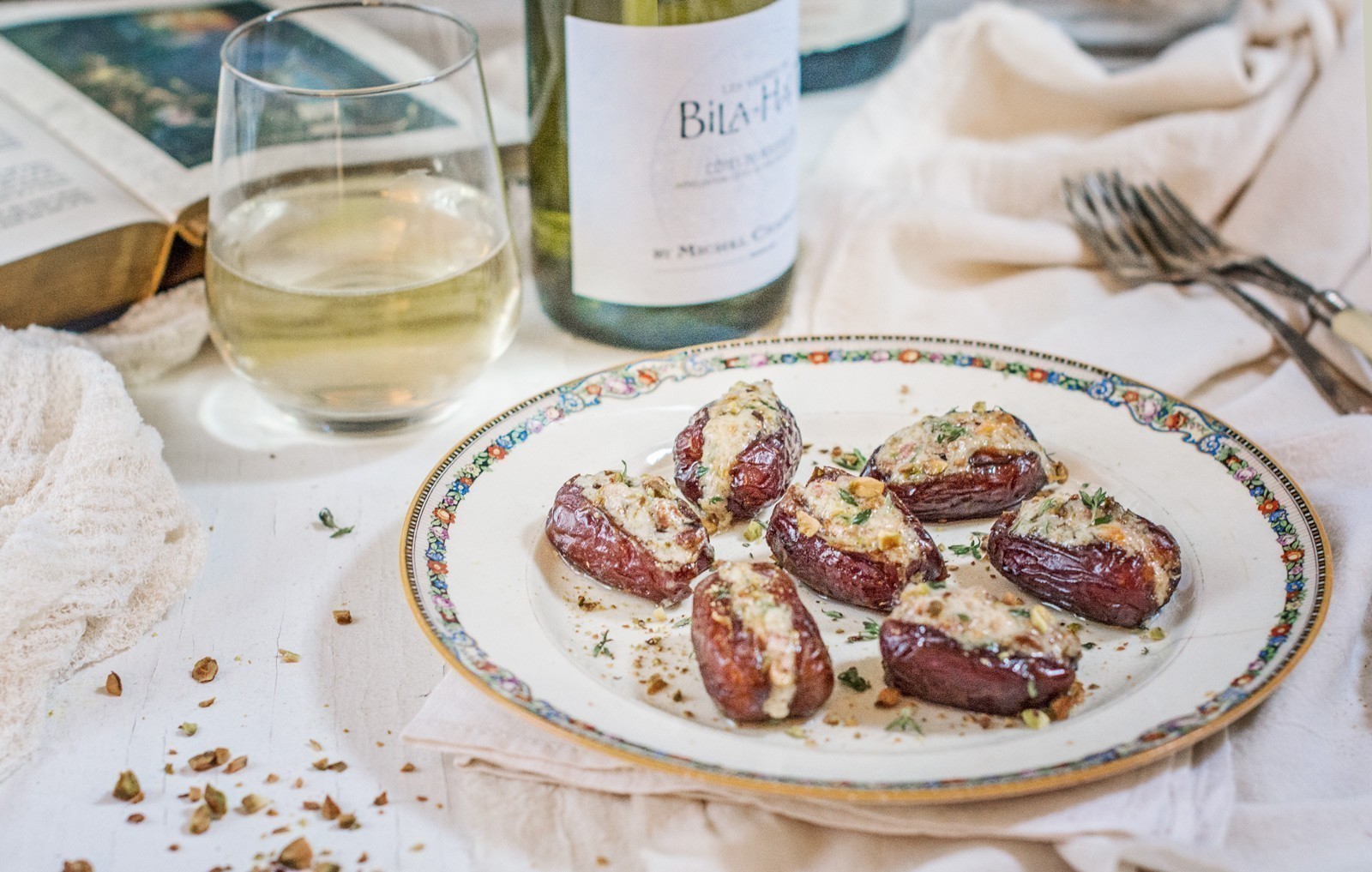 A decorative plate of stuffed dates, topped with herbs and cheese, rests on a white cloth beside a glass of white wine, a wine bottle, forks, and an open book. Crushed nuts are sprinkled around the stuffed dates for added texture.
