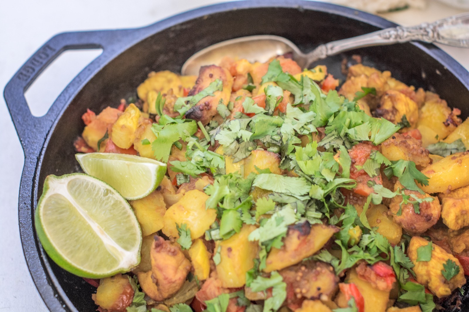 A cast iron skillet filled with a colorful potato chicken stew and vegetable stir-fry, garnished with fresh cilantro, served with lime wedges and a silver spoon.