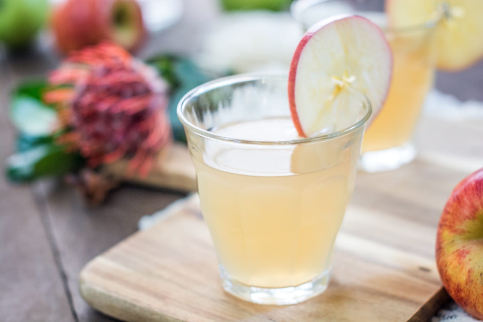 A glass of light-colored Apple Ginger Cocktail garnished with a thin apple slice sits on a wooden board, with fresh apples and blurred fruit in the background.