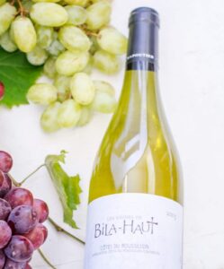 A bottle of Bila-Haut white wine lies on a white surface, surrounded by clusters of green and purple grapes, a green grape leaf, and some stuffed dates. The label reads Les Vignes de Bila-Haut, Michel Chapoutier.