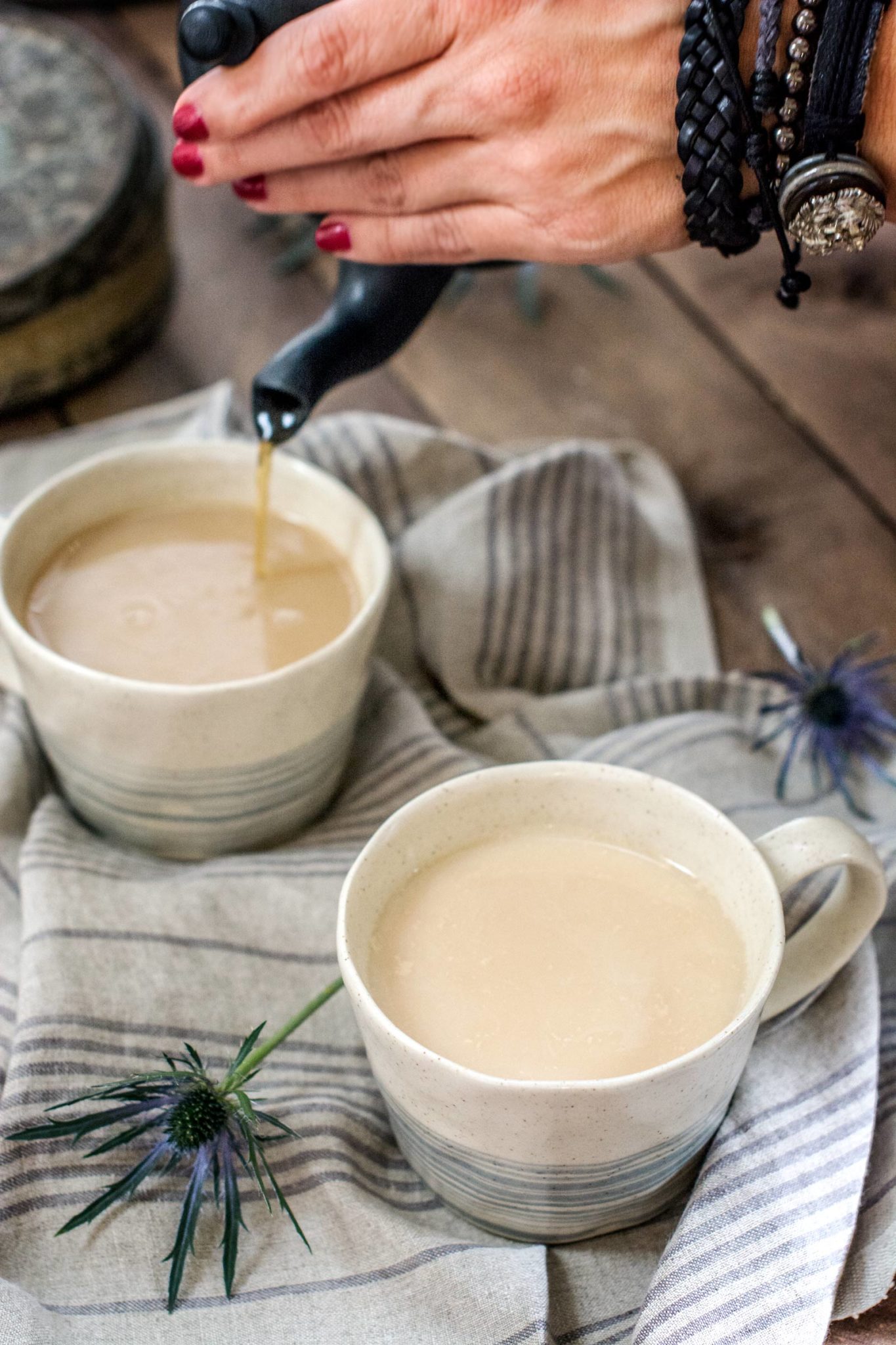 A hand pours Whiskey Chai into a cream-colored mug next to another filled mug, both placed on a striped cloth with decorative blue thistle flowers. The person has red nails and wears black bracelets and a watch.